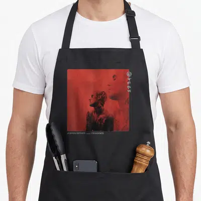 Justin novelty apron - Two male profiles on red background with bottom text JUSTIN BIEBER CHANGES — shown on a black cotton canvas apron against a white background