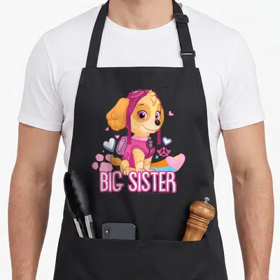 Skye sister novelty apron - Cartoon dog in pink hat surrounded by hearts and big sister text — shown on a black cotton canvas apron against a white background