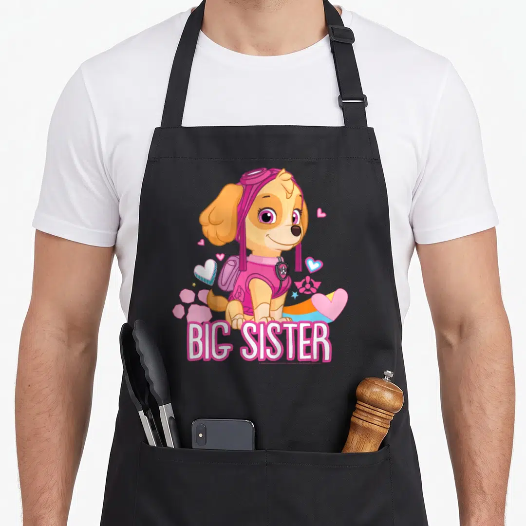 Skye sister novelty apron - Cartoon dog in pink hat surrounded by hearts and big sister text — shown on a black cotton canvas apron against a white background