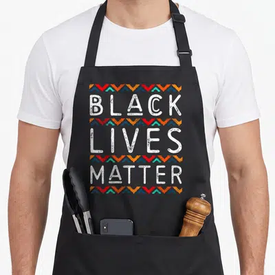 Pride african american aprons - White BLACK LIVES MATTER text separated by red orange teal and blue zig-zag pattern rows — shown on a black cotton canvas apron against a white background