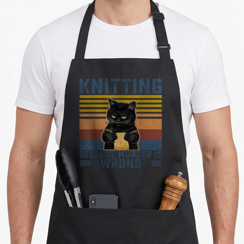 Murder knitting apron - Black cat knitting yellow swatch with blue text on striped background — shown on a black cotton canvas apron against a white background