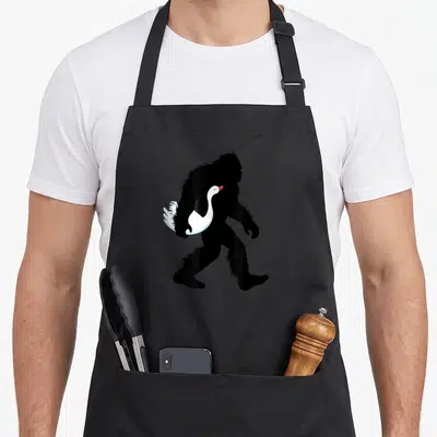 Bigfoot farming apron - Black Bigfoot silhouette walking right holding white swan with red beak — shown on a black cotton canvas apron against a white background