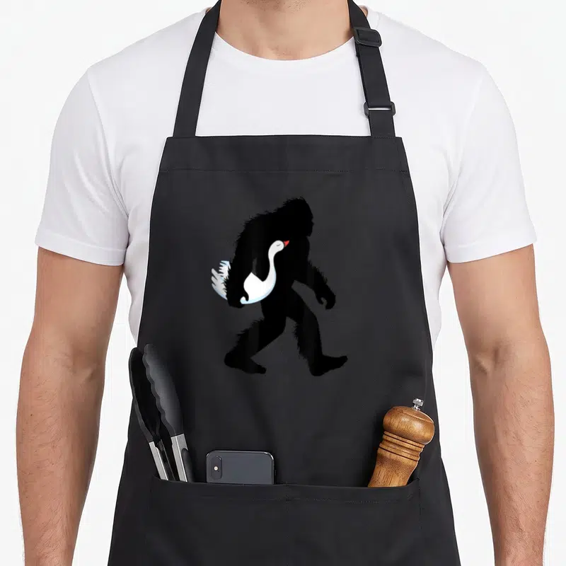 Bigfoot farming apron - Black Bigfoot silhouette walking right holding white swan with red beak — shown on a black cotton canvas apron against a white background