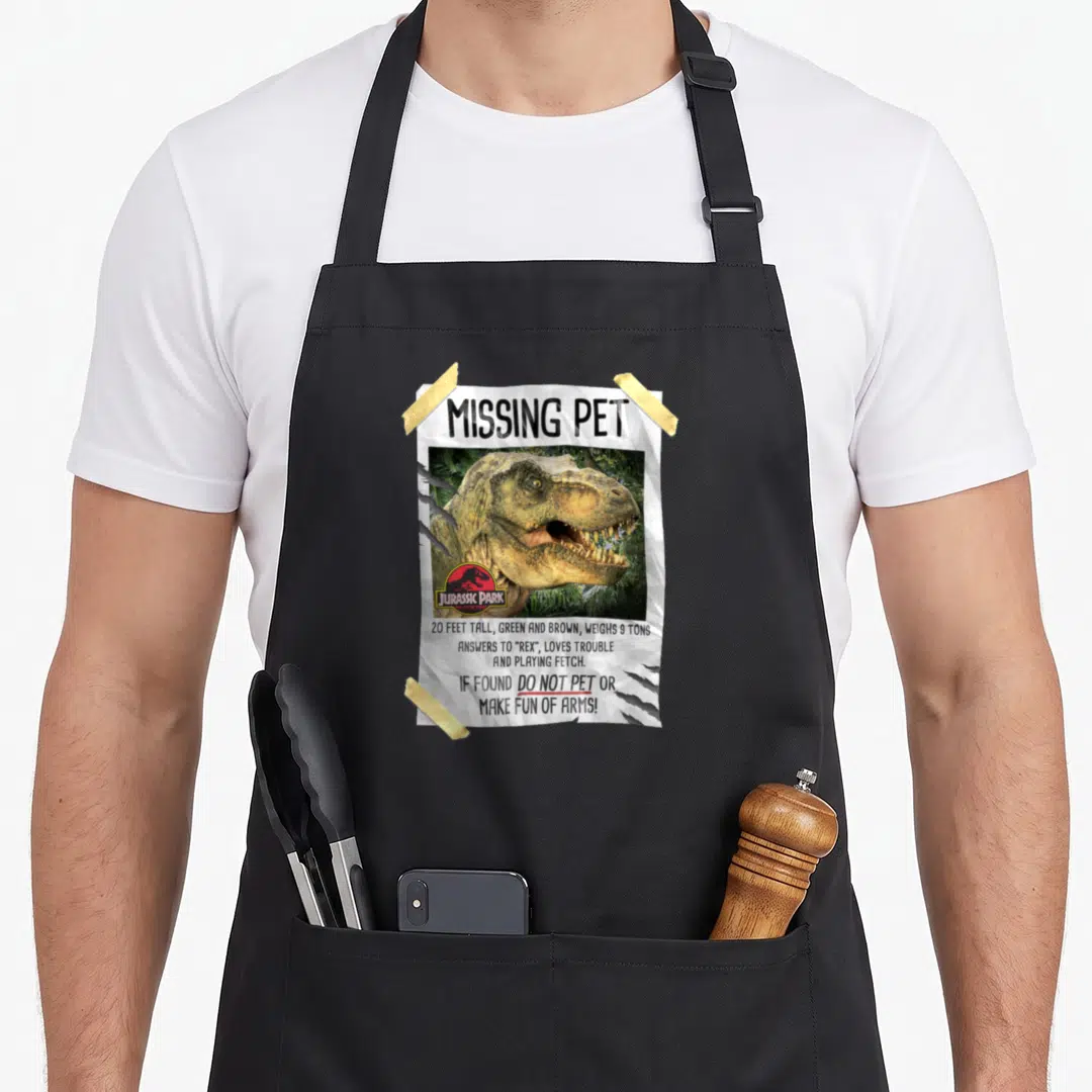 Missing pet apron - Taped missing pet flyer with T-Rex photo and Jurassic Park logo warning not to pet or make fun — shown on a black cotton canvas apron against a white background