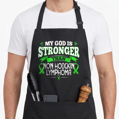 Non blood apron - White text MY GOD IS STRONGER THAN NON HODGKIN LYMPHOMA with green awareness ribbons — shown on a black cotton canvas apron against a white background