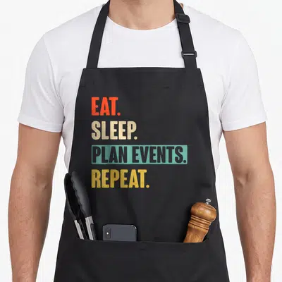 Eat event apron - Stacked text EAT SLEEP PLAN EVENTS REPEAT in orange beige teal yellow distressed font — shown on a black cotton canvas apron against a white background