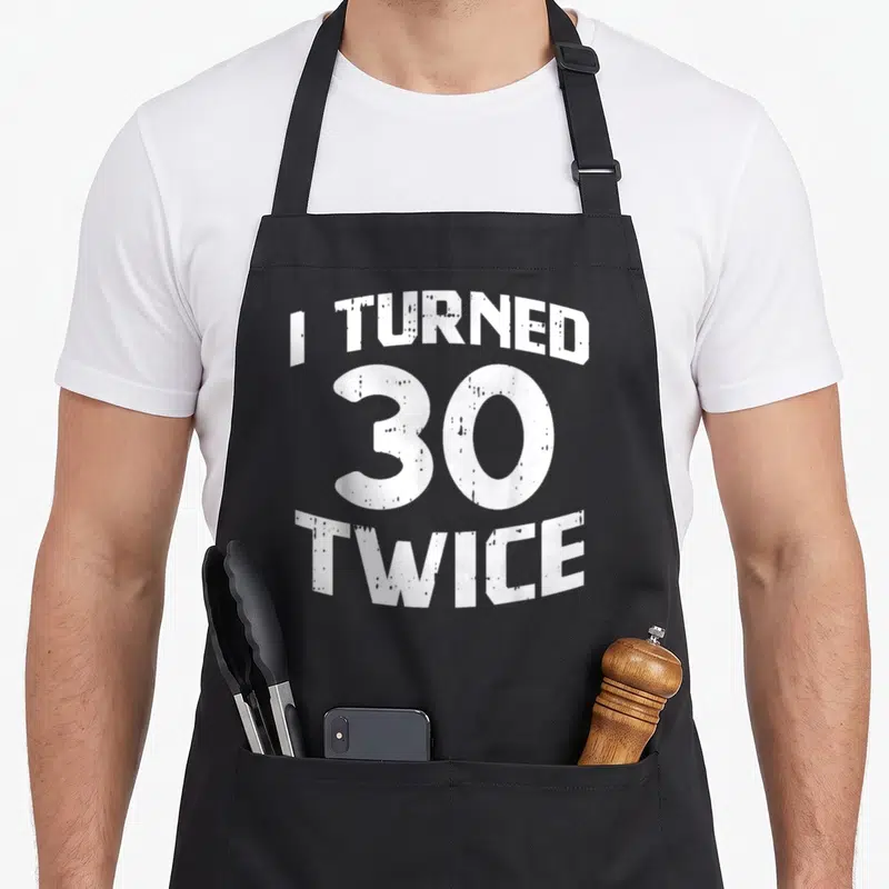 Boredkoalas 60th birthday apron - White distressed text reading I TURNED 30 TWICE in stacked arched layout — shown on a black cotton canvas apron against a white background