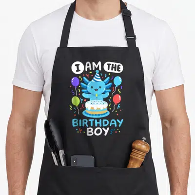 Birthday axolotl family aprons - Blue axolotl wearing party hat behind cake with I AM THE BIRTHDAY BOY text and balloons — shown on a black cotton canvas apron against a white background