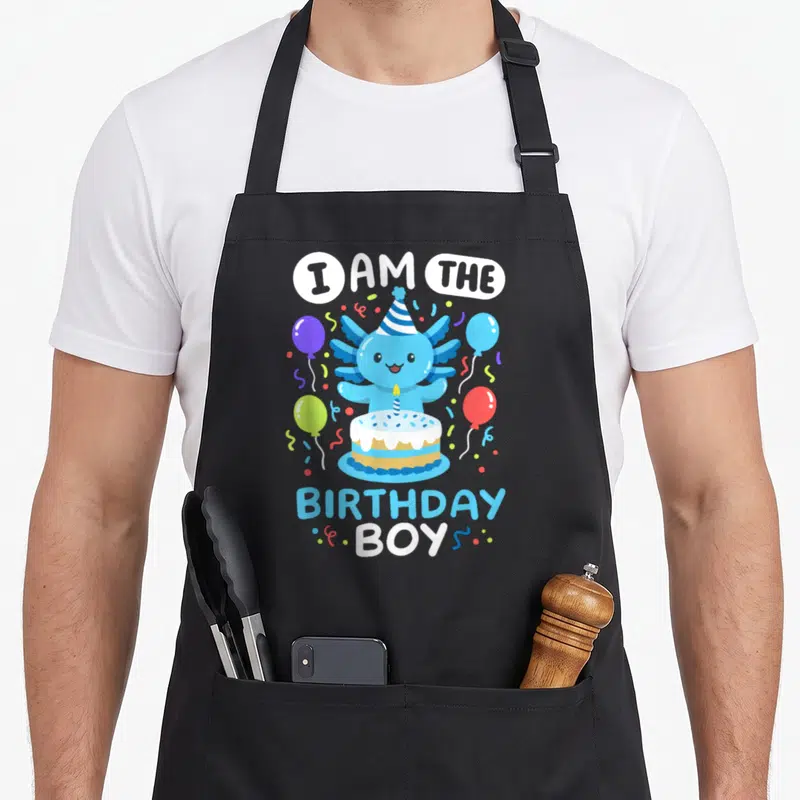 Birthday axolotl family aprons - Blue axolotl wearing party hat behind cake with I AM THE BIRTHDAY BOY text and balloons — shown on a black cotton canvas apron against a white background
