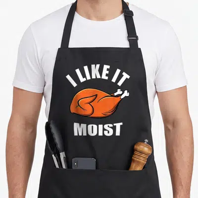 Funny moist thanksgiving apron - Cartoon roasted turkey with I LIKE IT MOIST text in orange and white — shown on a black cotton canvas apron against a white background