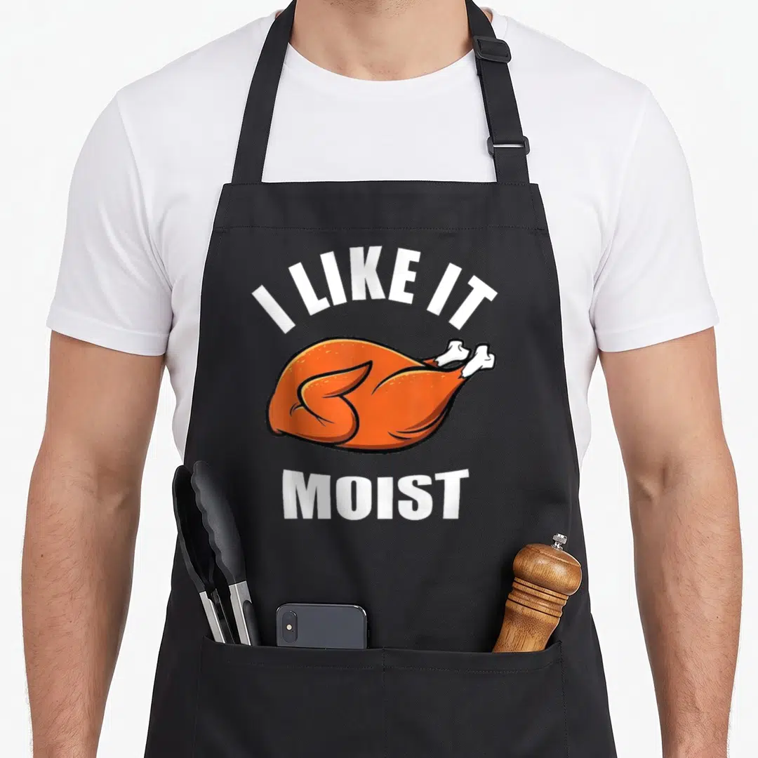 Funny moist thanksgiving apron - Cartoon roasted turkey with I LIKE IT MOIST text in orange and white — shown on a black cotton canvas apron against a white background