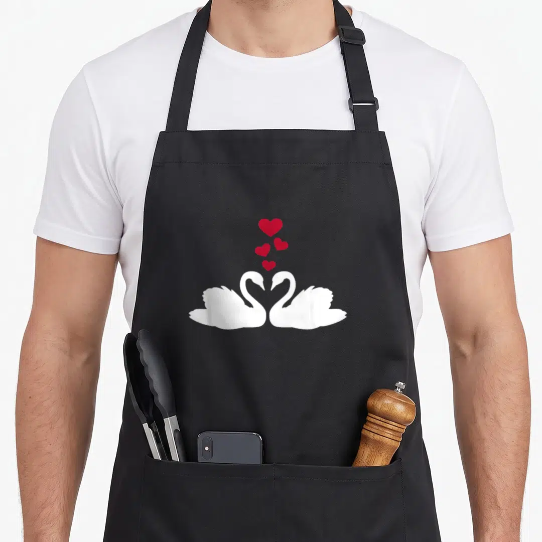 Funny i love my swan apron - Two white swans face each other with necks forming a heart, four red hearts float above — shown on a black cotton canvas apron against a white background