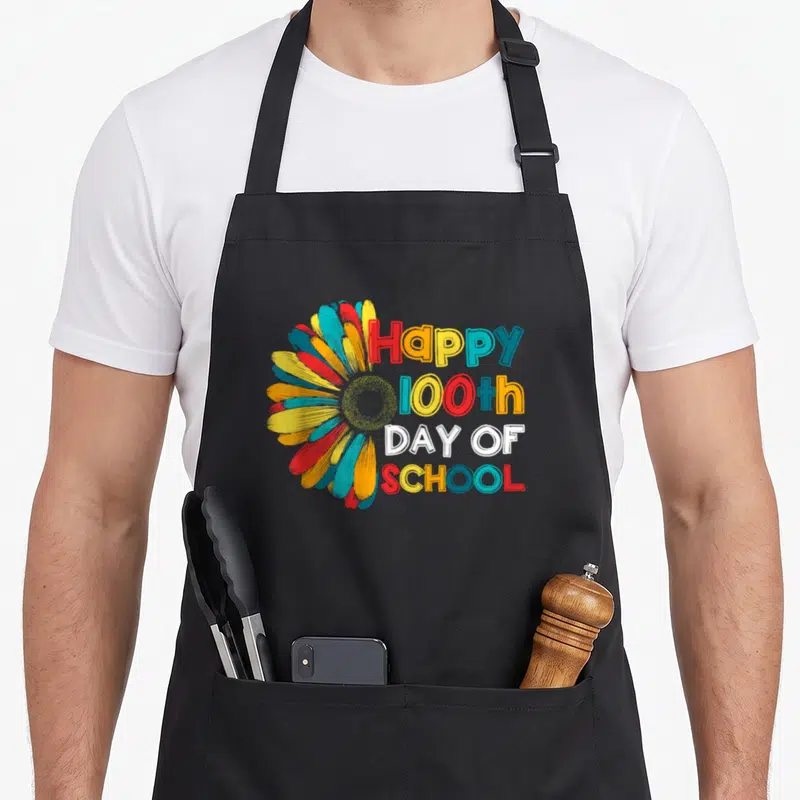 School kinder teacher apron - Multicolored flower and stacked Happy 100th Day of School text in red yellow blue white — shown on a black cotton canvas apron against a white background