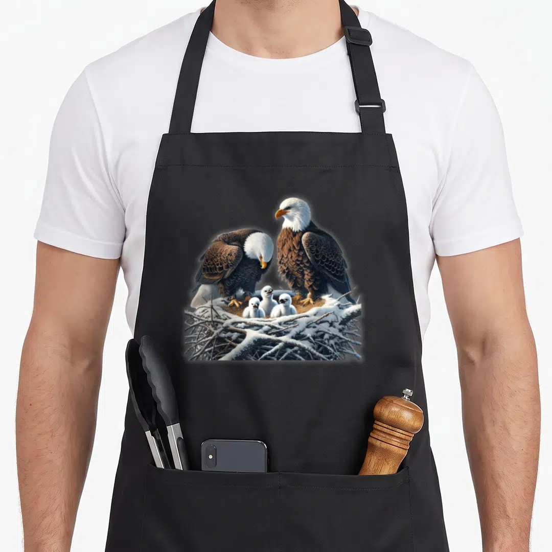 Three eagles apron - Two adult bald eagles and three white chicks in a snowy twig nest — shown on a black cotton canvas apron against a white background