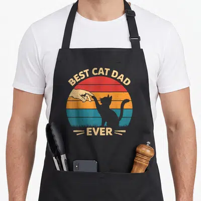 Cat owner father day apron - Retro striped sun with cat silhouette reaching for hand and best cat dad ever text — shown on a black cotton canvas apron against a white background