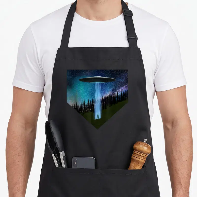 Saucer flying apron - UFO emitting blue beam to standing person in field below black trees inside shield shape — shown on a black cotton canvas apron against a white background