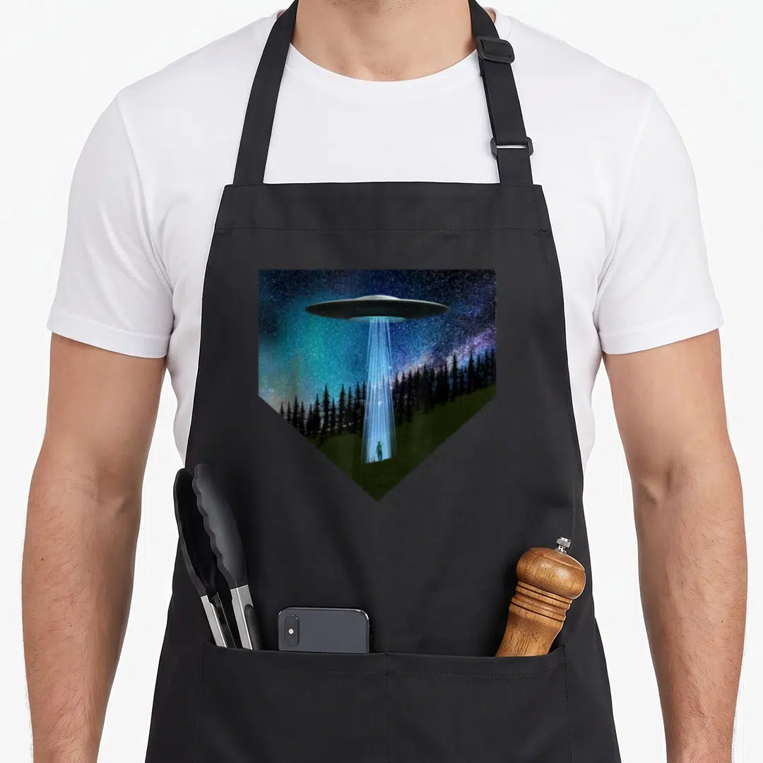Saucer flying apron - UFO emitting blue beam to standing person in field below black trees inside shield shape — shown on a black cotton canvas apron against a white background