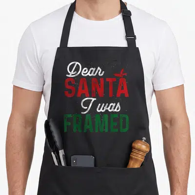 Santa womens christmas aprons - Distressed text reading Dear Santa I was Framed with red holly leaf accent in white red — shown on a black cotton canvas apron against a white background