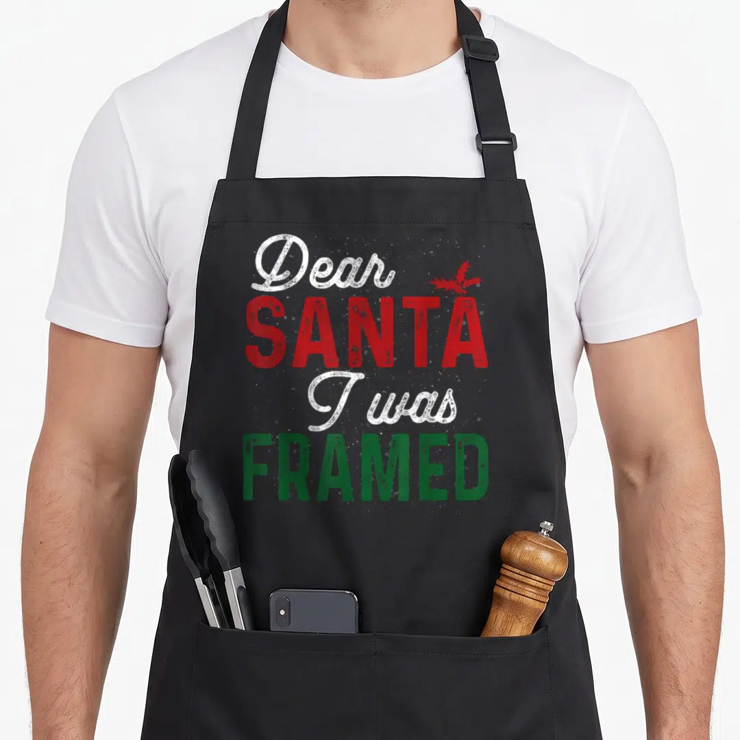 Santa womens christmas aprons - Distressed text reading Dear Santa I was Framed with red holly leaf accent in white red — shown on a black cotton canvas apron against a white background
