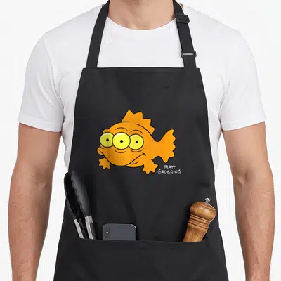 Blinky fish apron - Three-eyed orange cartoon fish with yellow eyes and Matt Groening signature text — shown on a black cotton canvas apron against a white background