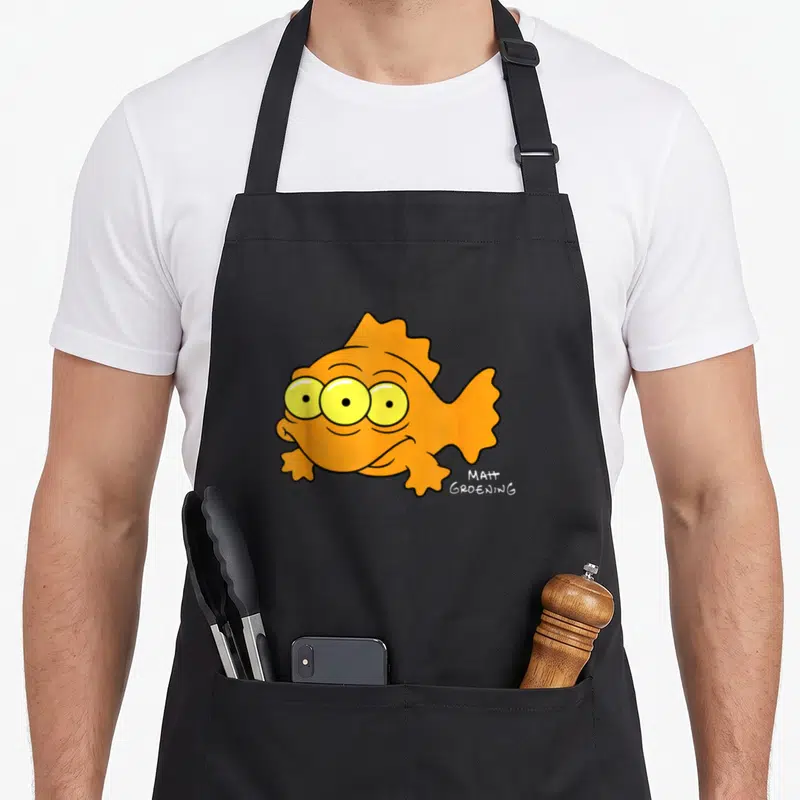 Blinky fish apron - Three-eyed orange cartoon fish with yellow eyes and Matt Groening signature text — shown on a black cotton canvas apron against a white background
