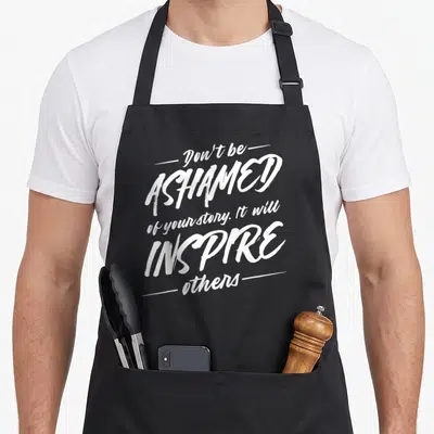 Drug recovery day apron - White stacked typography reading Don't be ASHAMED of your story on black fabric — shown on a black cotton canvas apron against a white background
