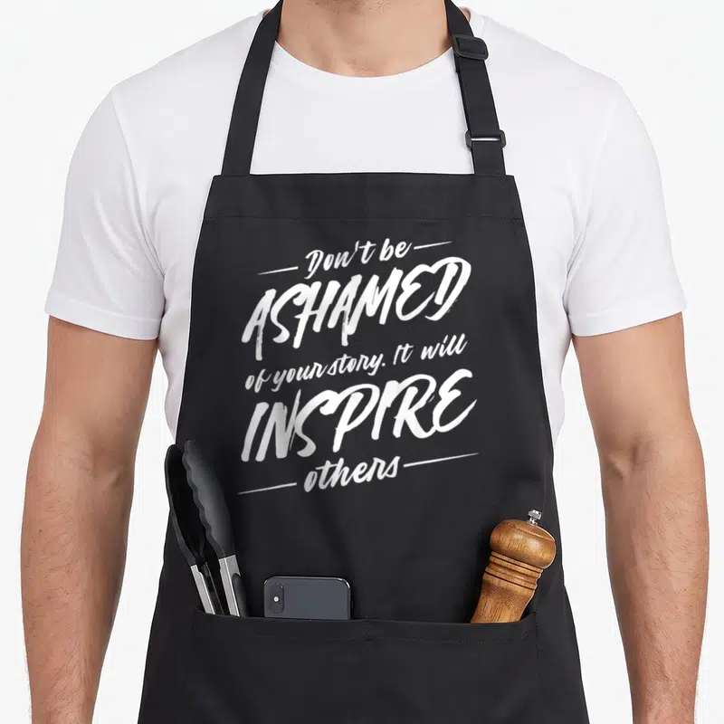 Drug recovery day apron - White stacked typography reading Don't be ASHAMED of your story on black fabric — shown on a black cotton canvas apron against a white background