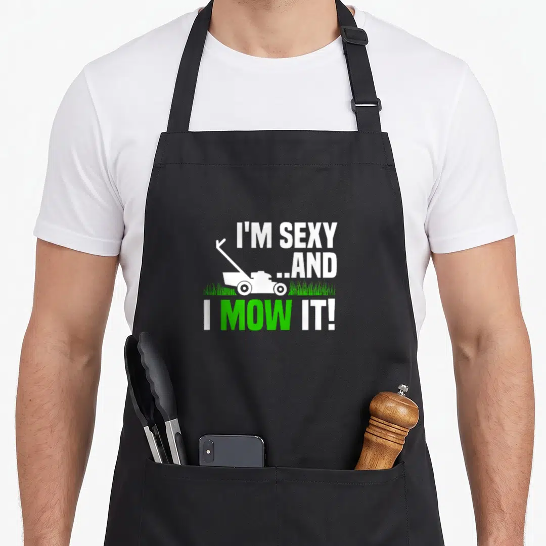 Mow mowing lawn apron - Stacked white text I'M SEXY I MOW IT with mower on green grass — shown on a black cotton canvas apron against a white background