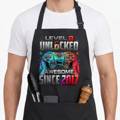 Unlocked 8th gaming aprons - Colorful game controller with LEVEL 8 UNLOCKED AWESOME SINCE 2017 text and stars — shown on a black cotton canvas apron against a white background