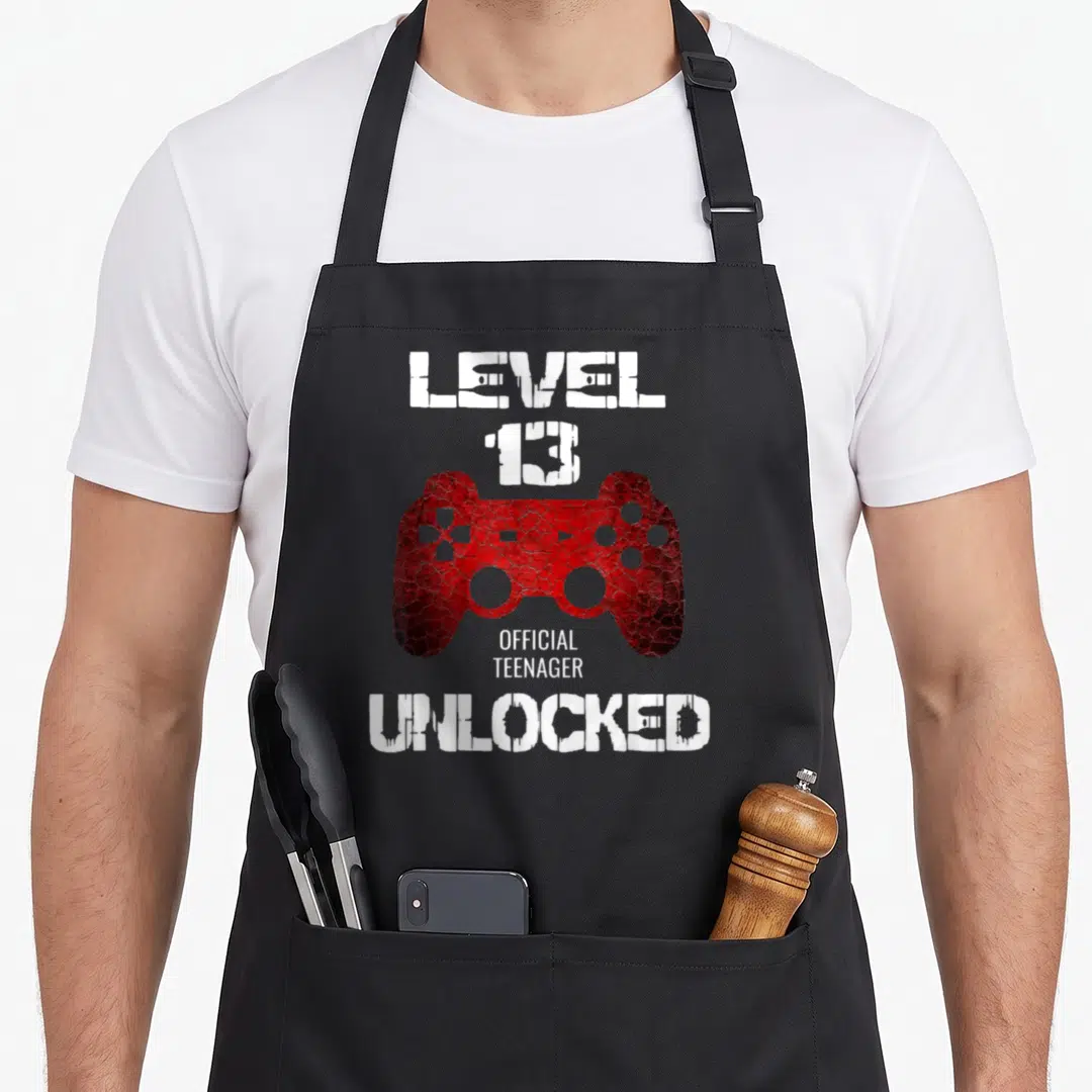 Level unlocked 13th birthday apron - Distressed red game controller with white text reading LEVEL 13 OFFICIAL TEENAGER — shown on a black cotton canvas apron against a white background