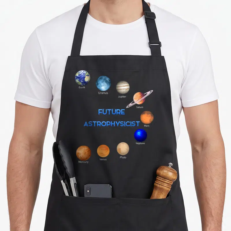 Future astronomy lover apron - Nine labeled planets orbiting FUTURE ASTROPHYSICIST text in blue orange brown white — shown on a black cotton canvas apron against a white background