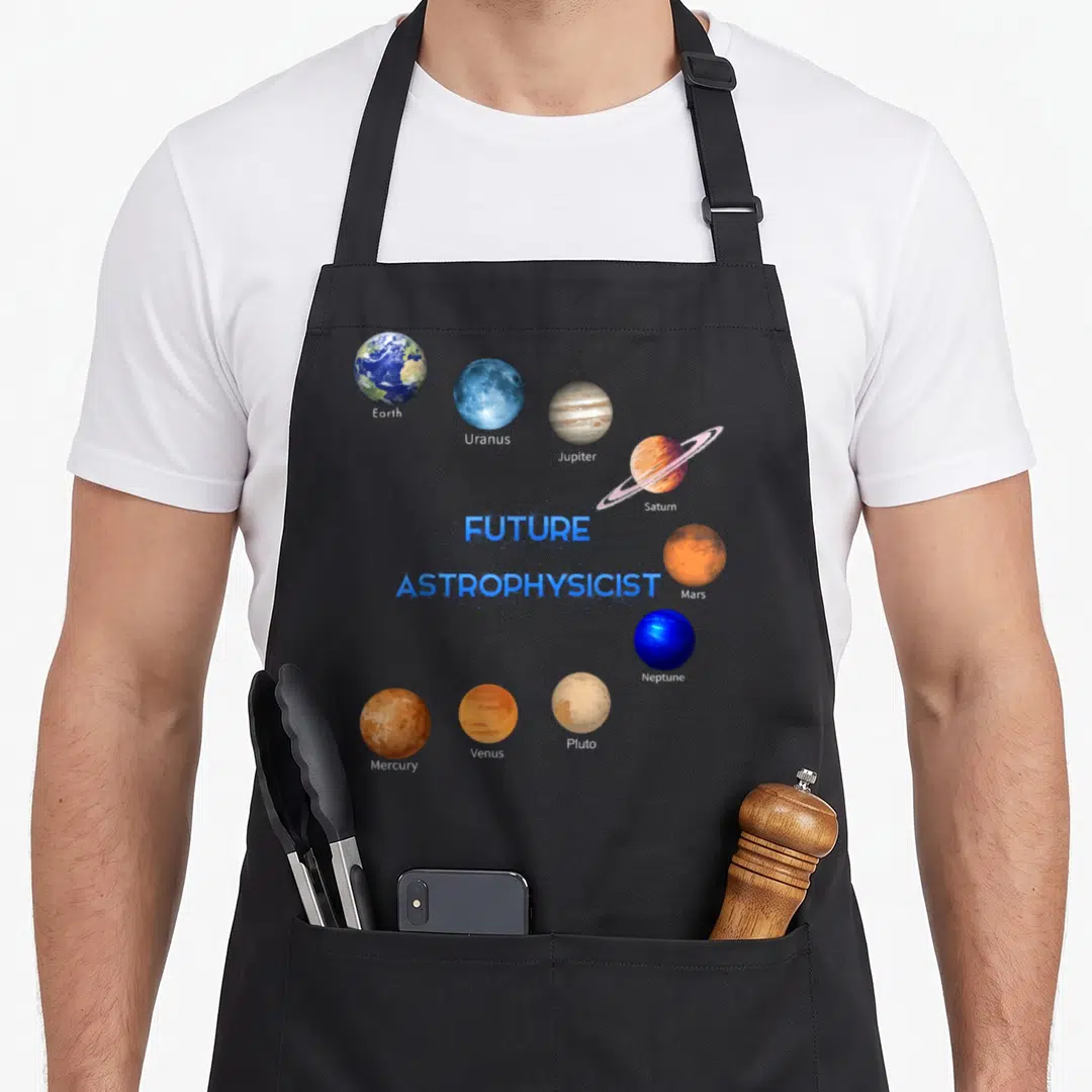 Future astronomy lover apron - Nine labeled planets orbiting FUTURE ASTROPHYSICIST text in blue orange brown white — shown on a black cotton canvas apron against a white background