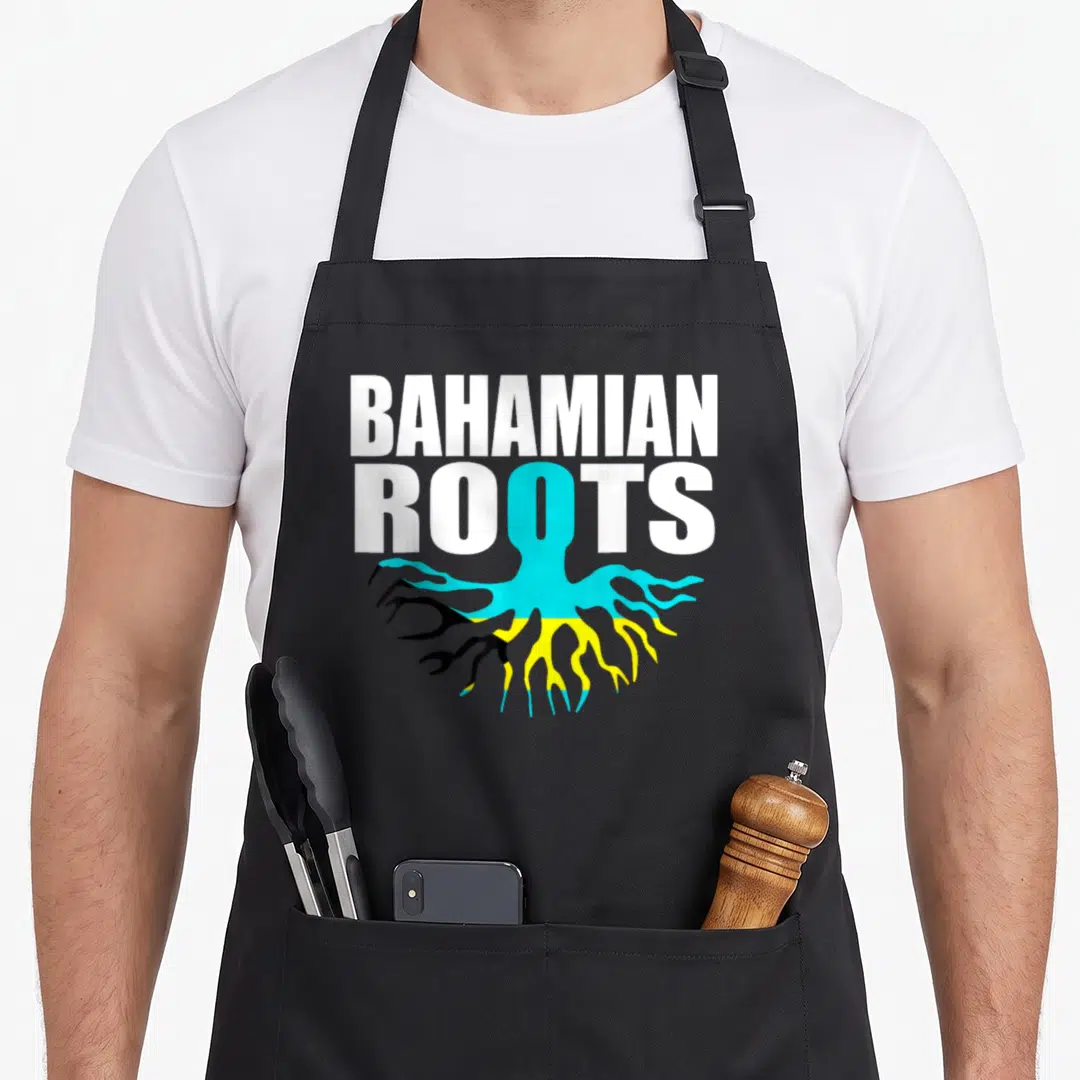 Flagcastle pride apron - BAHAMIAN ROOTS text with blue yellow and black flag-colored roots extending below — shown on a black cotton canvas apron against a white background