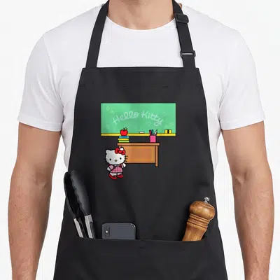 Teacher classroom aprons - Hello Kitty cartoon standing by brown desk with green chalkboard and red apple — shown on a black cotton canvas apron against a white background