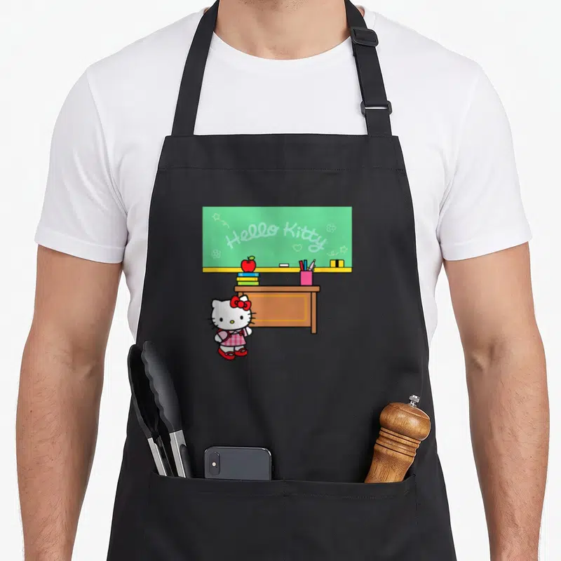 Teacher classroom aprons - Hello Kitty cartoon standing by brown desk with green chalkboard and red apple — shown on a black cotton canvas apron against a white background
