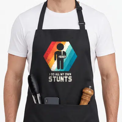 Arm recovering apron - Grey figure in sling inside striped hexagon with text I DO ALL MY OWN STUNTS — shown on a black cotton canvas apron against a white background