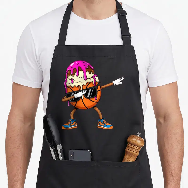 Dabbing ice cream apron - Cartoon orange basketball character with sunglasses and ice cream scoop doing a dab pose — shown on a black cotton canvas apron against a white background