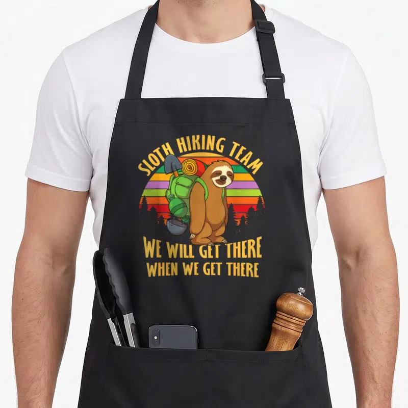 Cool hiking sloth apron - Cartoon sloth with backpack under SLOTH HIKING TEAM text and striped sun — shown on a black cotton canvas apron against a white background