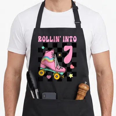 Roller skating 7th birthday apron - Rollin' Into text above pink skate and number 7 on checkerboard with stars — shown on a black cotton canvas apron against a white background