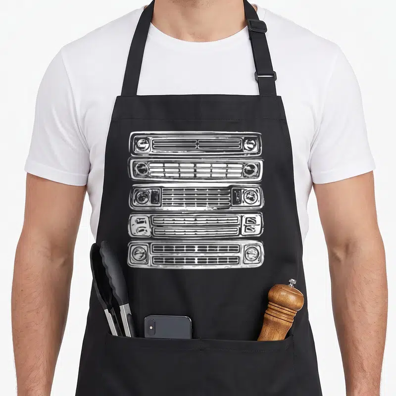 Funny truck apron - Five white car grilles with headlights stacked vertically in line art style — shown on a black cotton canvas apron against a white background