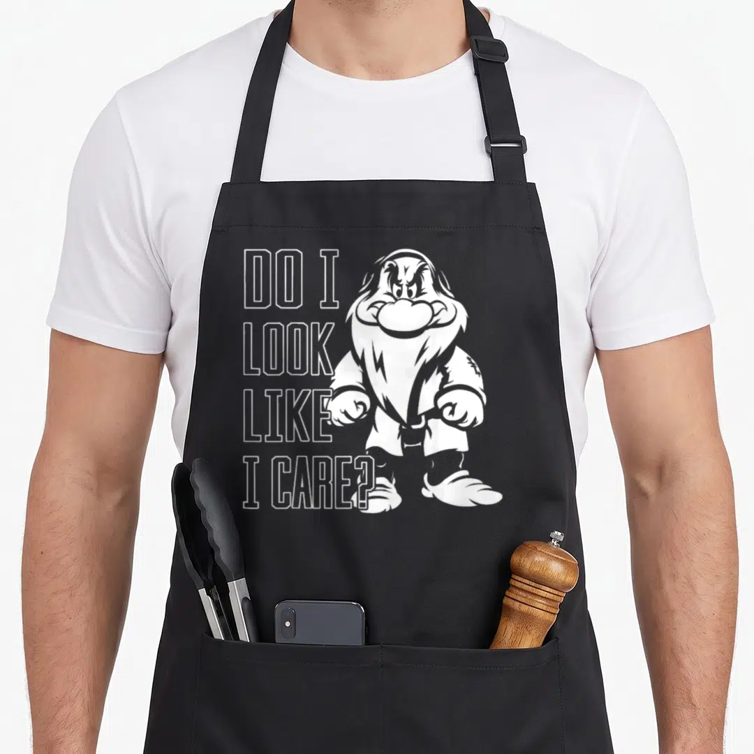 Care snow white apron - Grumpy dwarf illustration next to stacked text DO I LOOK LIKE I CARE in white outline — shown on a black cotton canvas apron against a white background