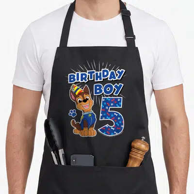 Chase age birthday apron - Cartoon dog with party hat next to number 5 filled with paw prints and bones — shown on a black cotton canvas apron against a white background