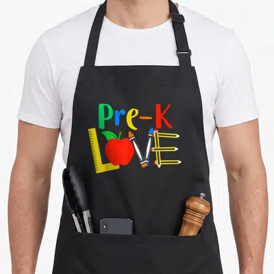 Classroom teacher apron - Pre-K text above LOVE formed by ruler, apple, and pencils in primary colors — shown on a black cotton canvas apron against a white background