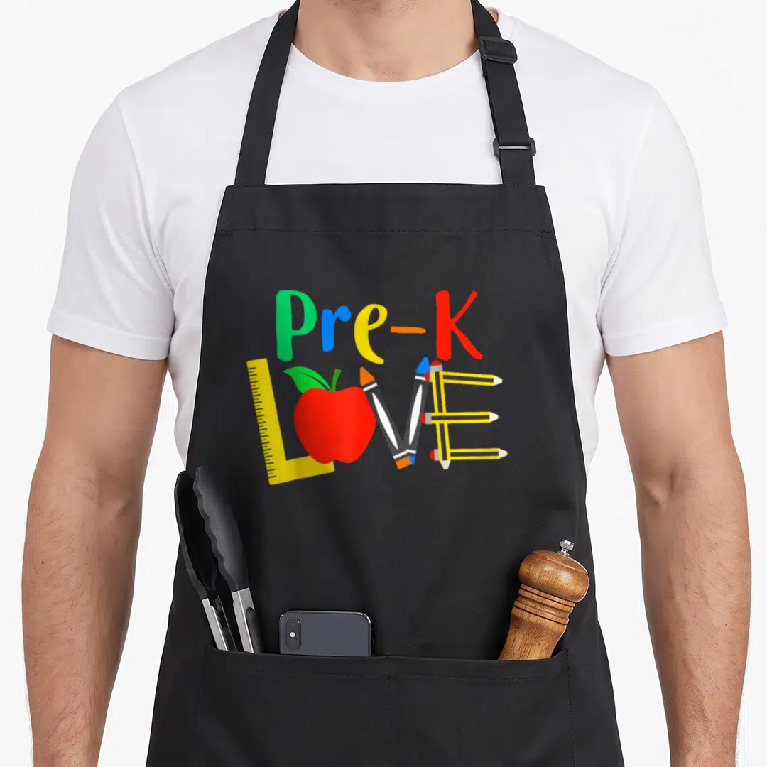 Classroom teacher apron - Pre-K text above LOVE formed by ruler, apple, and pencils in primary colors — shown on a black cotton canvas apron against a white background