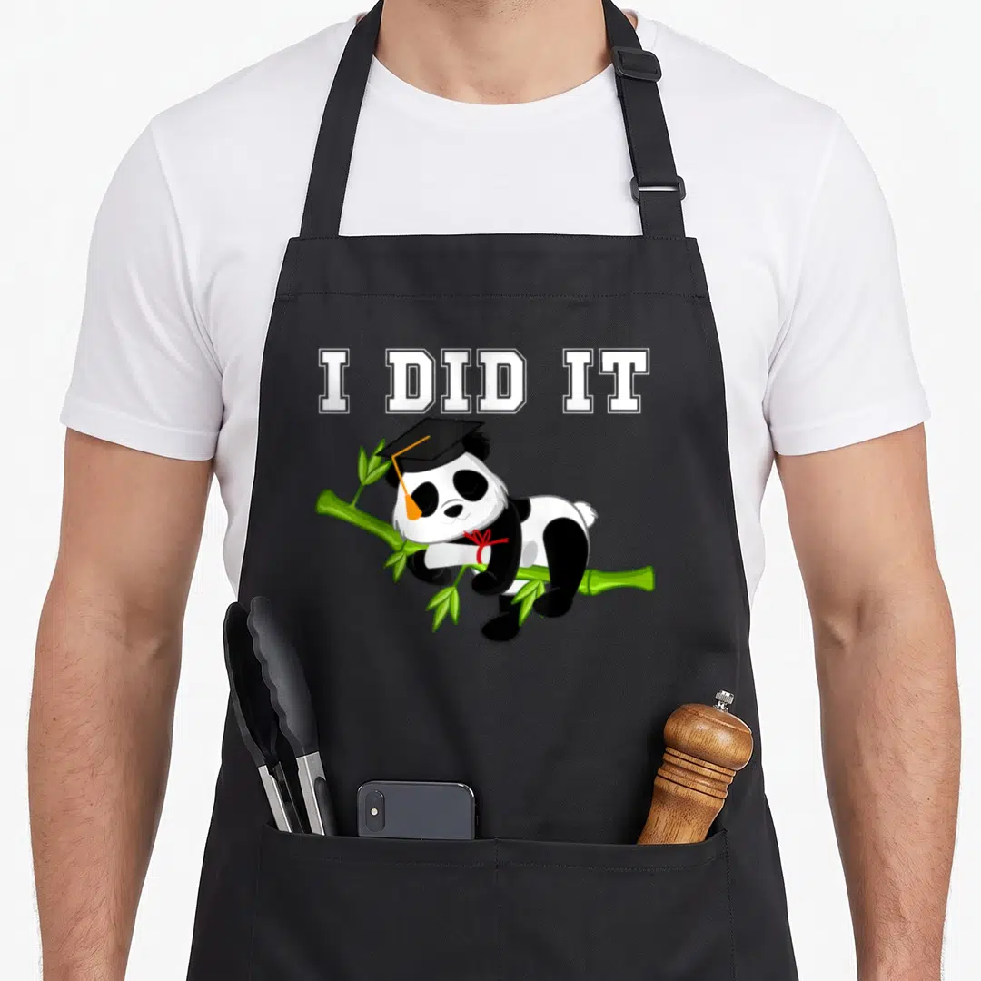 Graduation kawaii apron - I DID IT text above a panda in a cap lying on bamboo holding a scroll — shown on a black cotton canvas apron against a white background