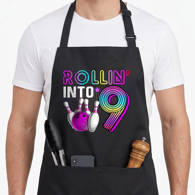 Bowling 9th birthday apron - Rollin into 9 text with rainbow number 9 and bowling ball pins — shown on a black cotton canvas apron against a white background