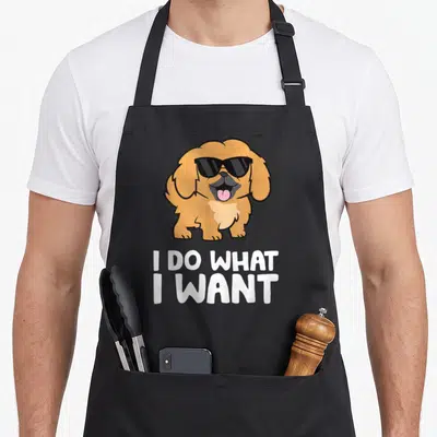 Cool pekingese dog apron - Cartoon tan Pekingese dog wearing black sunglasses above white text I DO WHAT I WANT — shown on a black cotton canvas apron against a white background