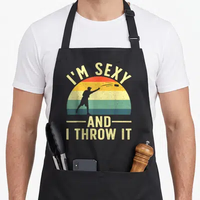 Corn game aprons - Retro disc thrower silhouette inside colorful sunset arch with text I'M SEXY AND I THROW IT — shown on a black cotton canvas apron against a white background