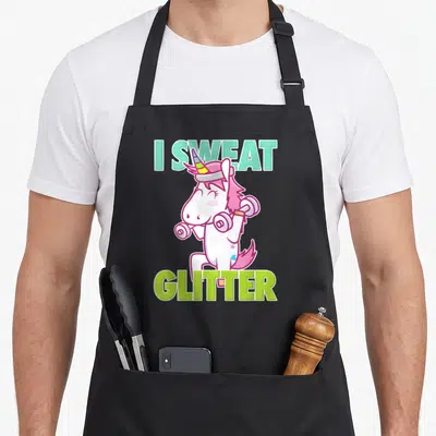 Sweat sassy apron - I Sweat Glitter text framing white unicorn lifting two dumbbells in mint and lime green — shown on a black cotton canvas apron against a white background