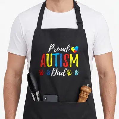 Autism family aprons - Proud AUTISM Dad text with puzzle heart and four colorful handprints in red yellow blue and white — shown on a black cotton canvas apron against a white background